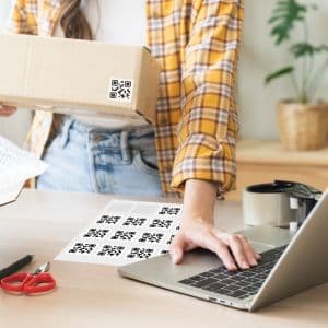 A person holding a box with one hand on their laptop keyboard, QR code stickers are laid out on the table